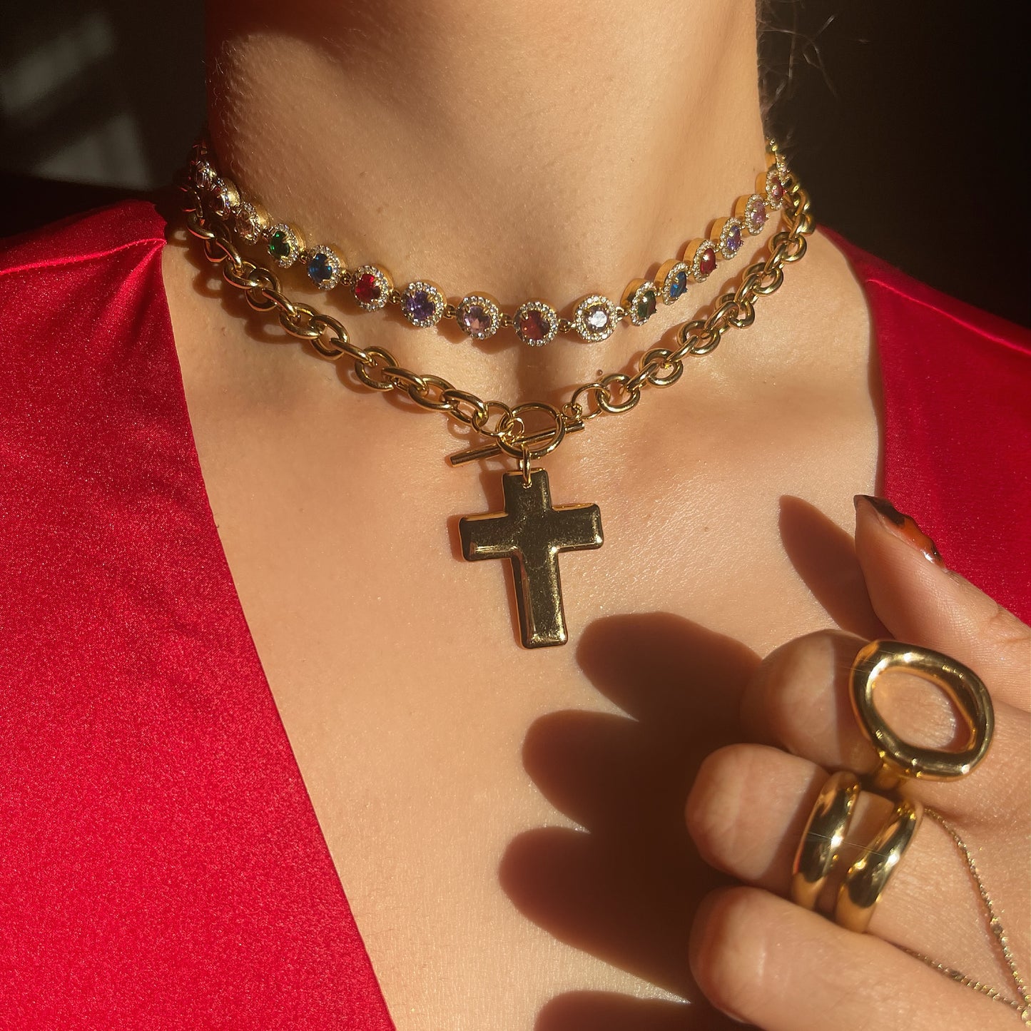 Close-up of a person wearing a gold necklace with a cross pendant and multiple rings on a red background.