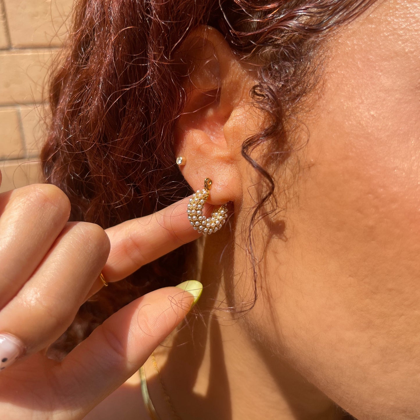 Close-up of an ear with a pearl gold hoop earring, held by a hand against a blurred background.