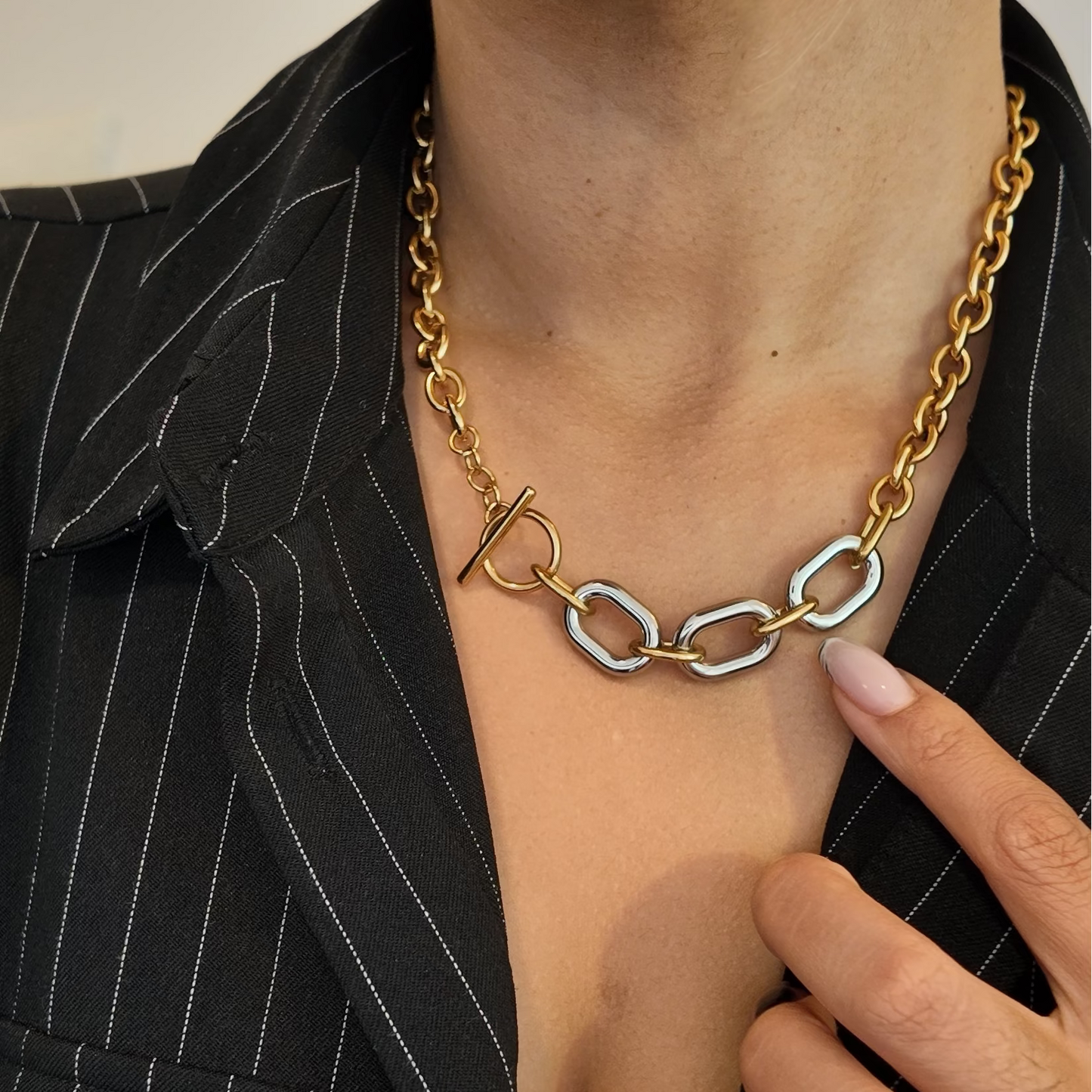 A close-up of a person wearing a gold chain necklace with a distinctive interlocking link design and a toggle clasp, styled over a black pinstripe blazer.