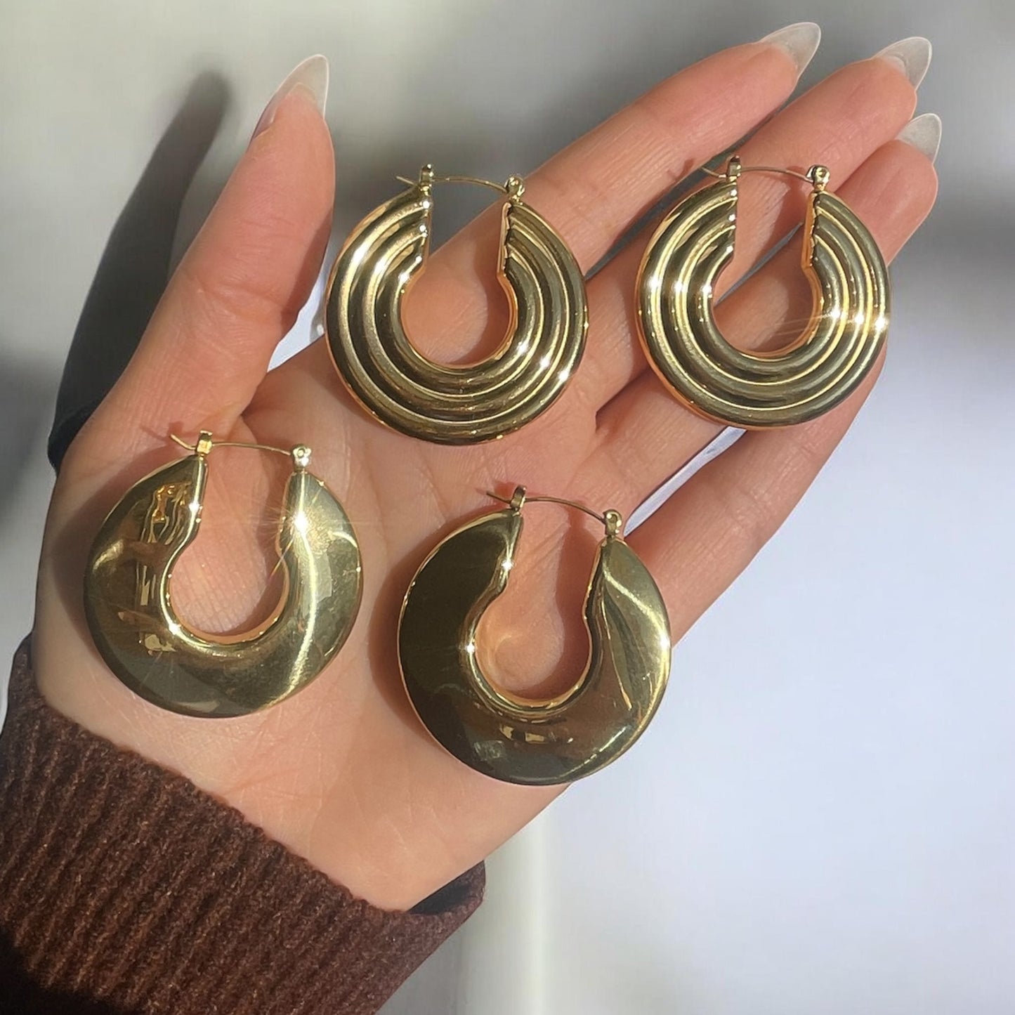 Gold hoop earrings held in a hand against a neutral background