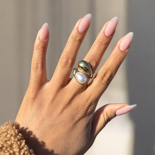 Hand wearing a gold ring with a pearl-like stone against a neutral background