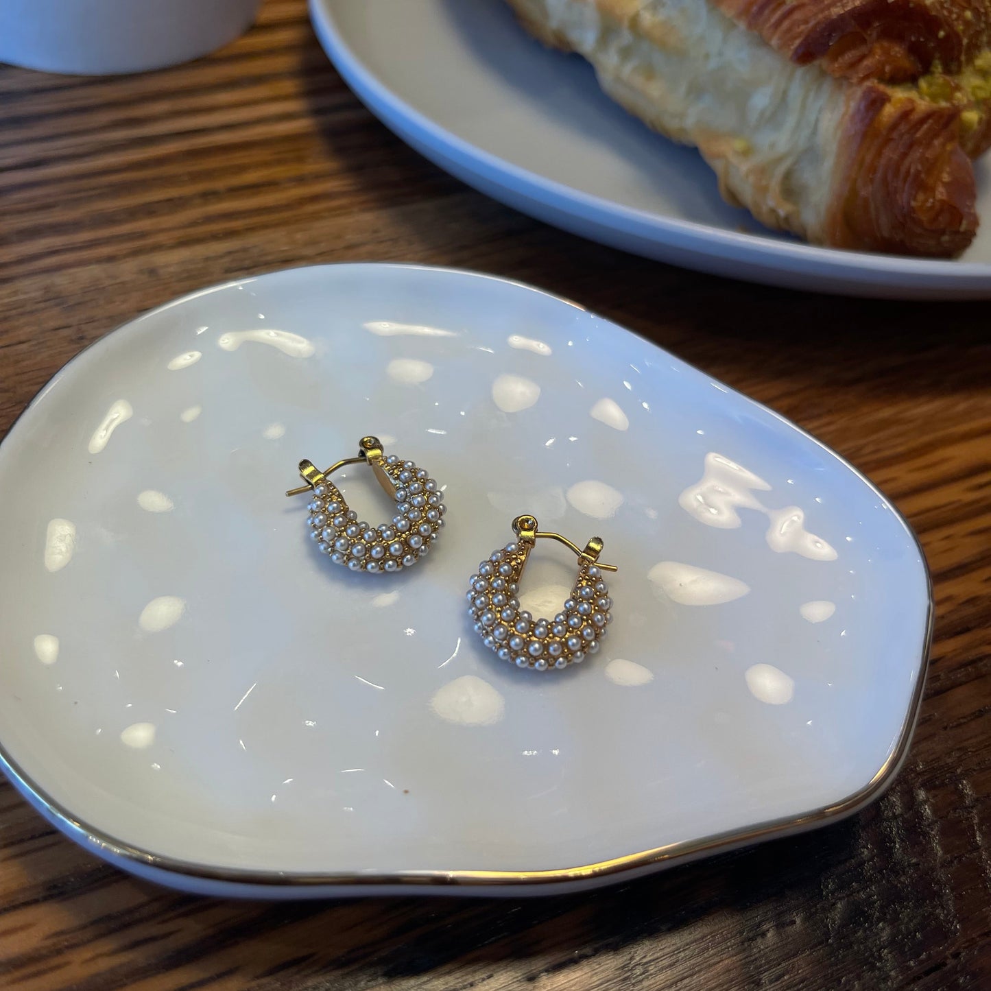Jewelry box with pearl huggie hoop earrings on a wooden table next to a pastry.