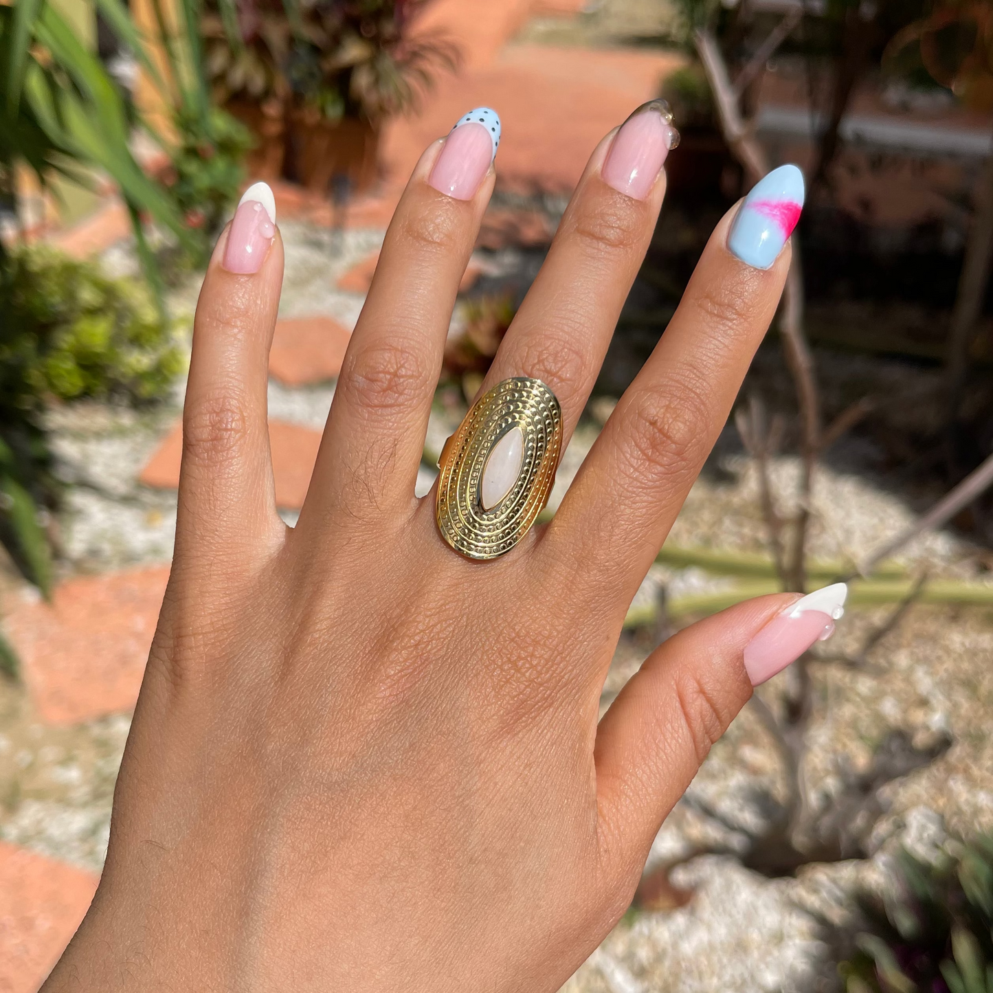 Hand wearing a gold ring with colorful nail polish against a garden background