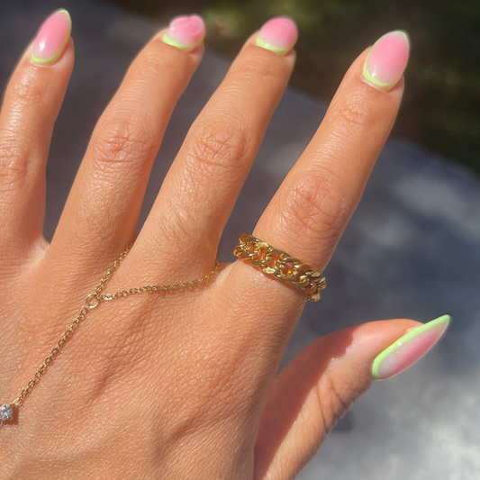 Hand with pink and green nail polish wearing a gold ring and hand chain, blurred background