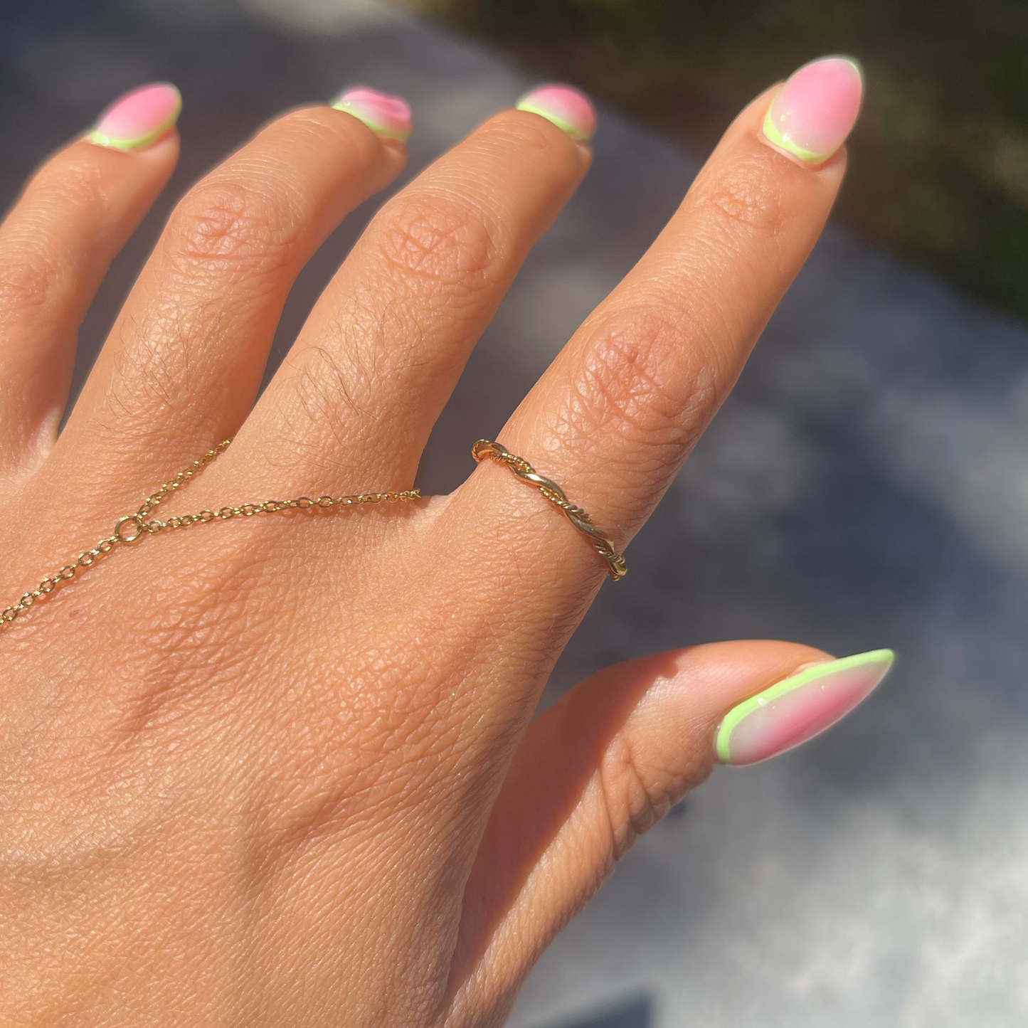 Hand with pink and green nail polish wearing a gold ring and bracelet against a blurred outdoor background