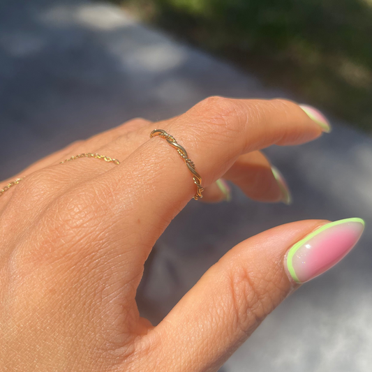 Hand with green and pink nail polish wearing a gold ring against a blurred outdoor background