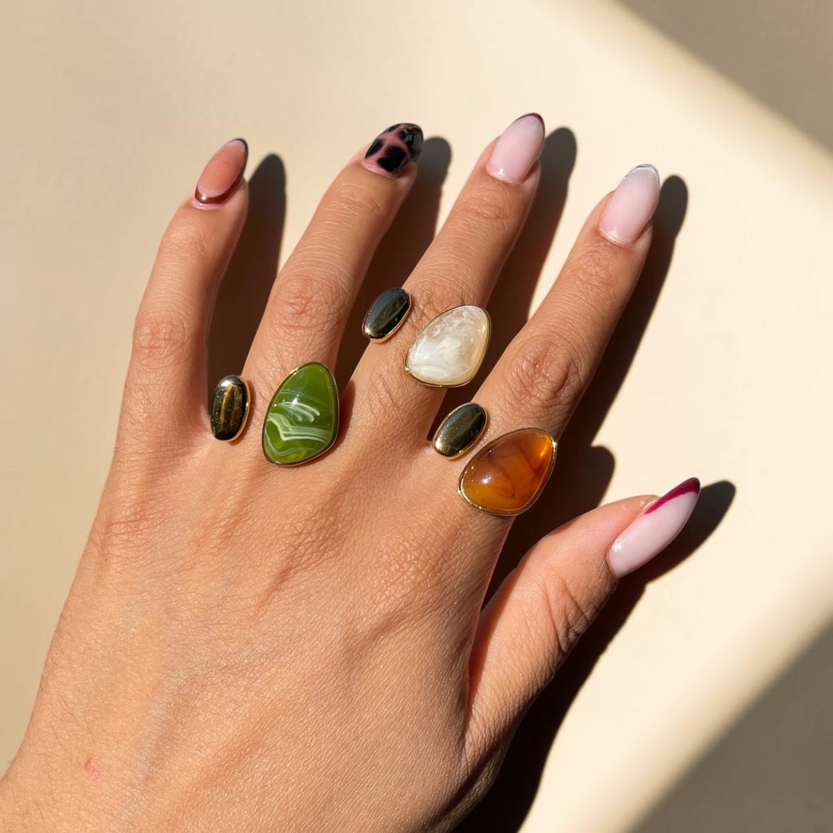 A hand with manicured nails wearing five distinct natural stone rings, photographed in bright sunlight against a warm beige background.