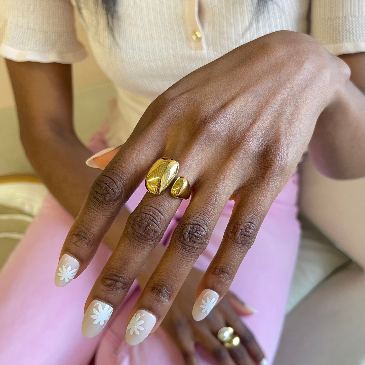 Close-up of a hand with gold rings and light-colored nail polish, wearing a beige sweater and pink pants.