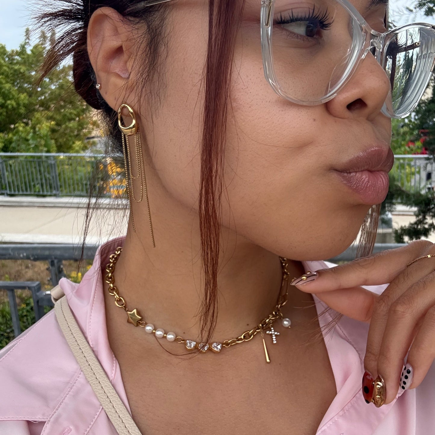 Person wearing glasses, a gold pearl necklace, and a pink shirt with a blurred outdoor background