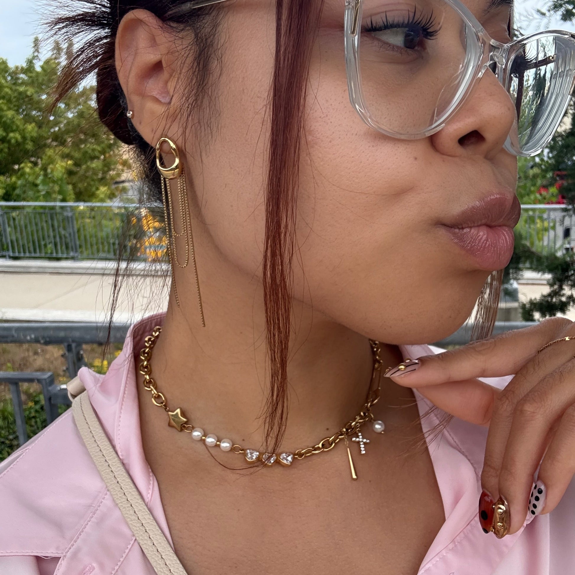 Person wearing glasses, a gold pearl necklace, and a pink shirt with a blurred outdoor background