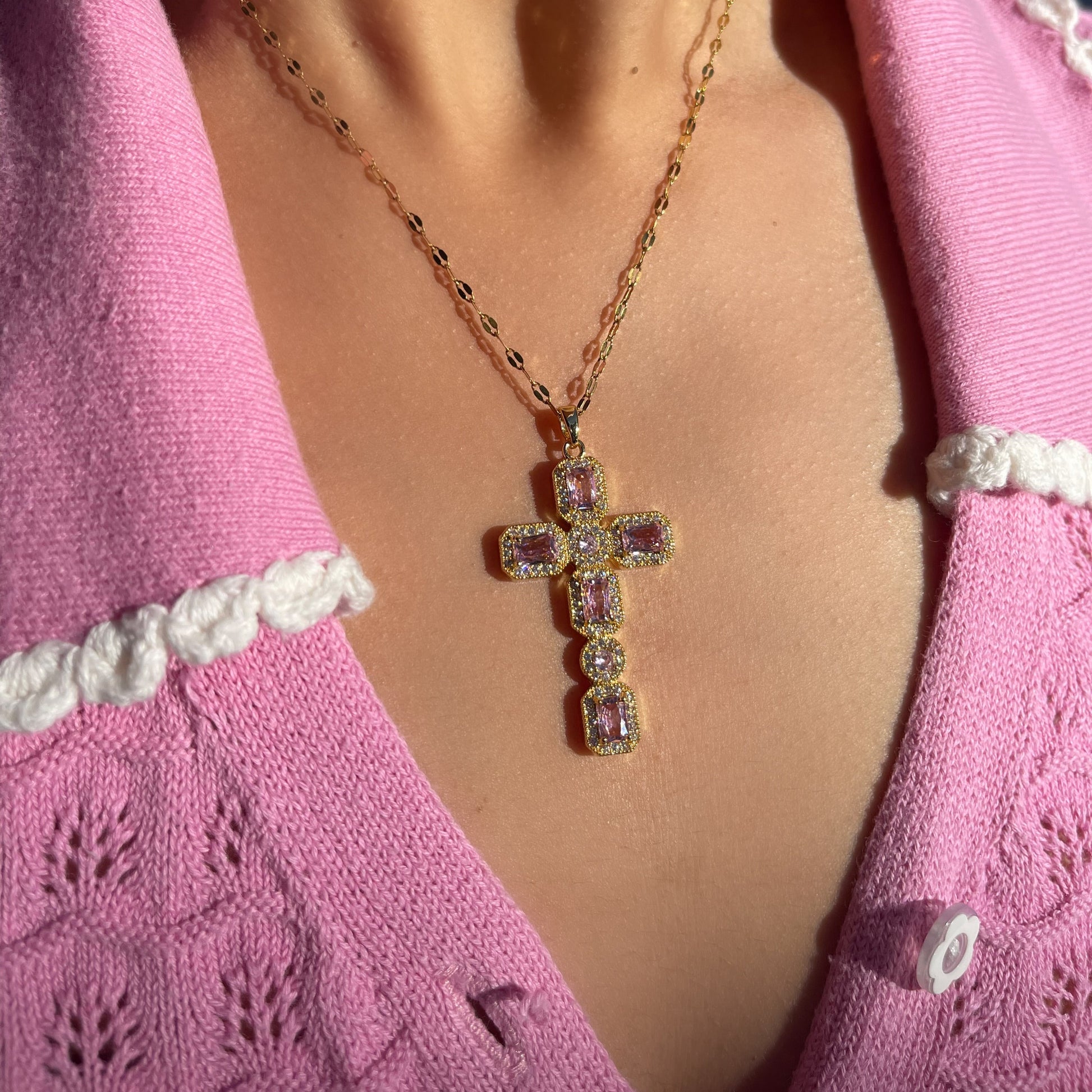 Gold cross necklace with pink gemstones worn by a person in a pink sweater.