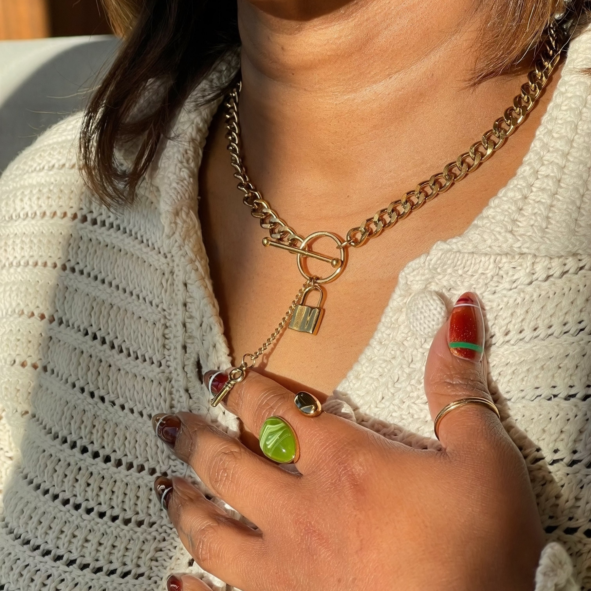 A close-up of a person wearing a gold chain necklace with a toggle clasp and a small padlock charm, styled with a white knit top and colorful nail polish.