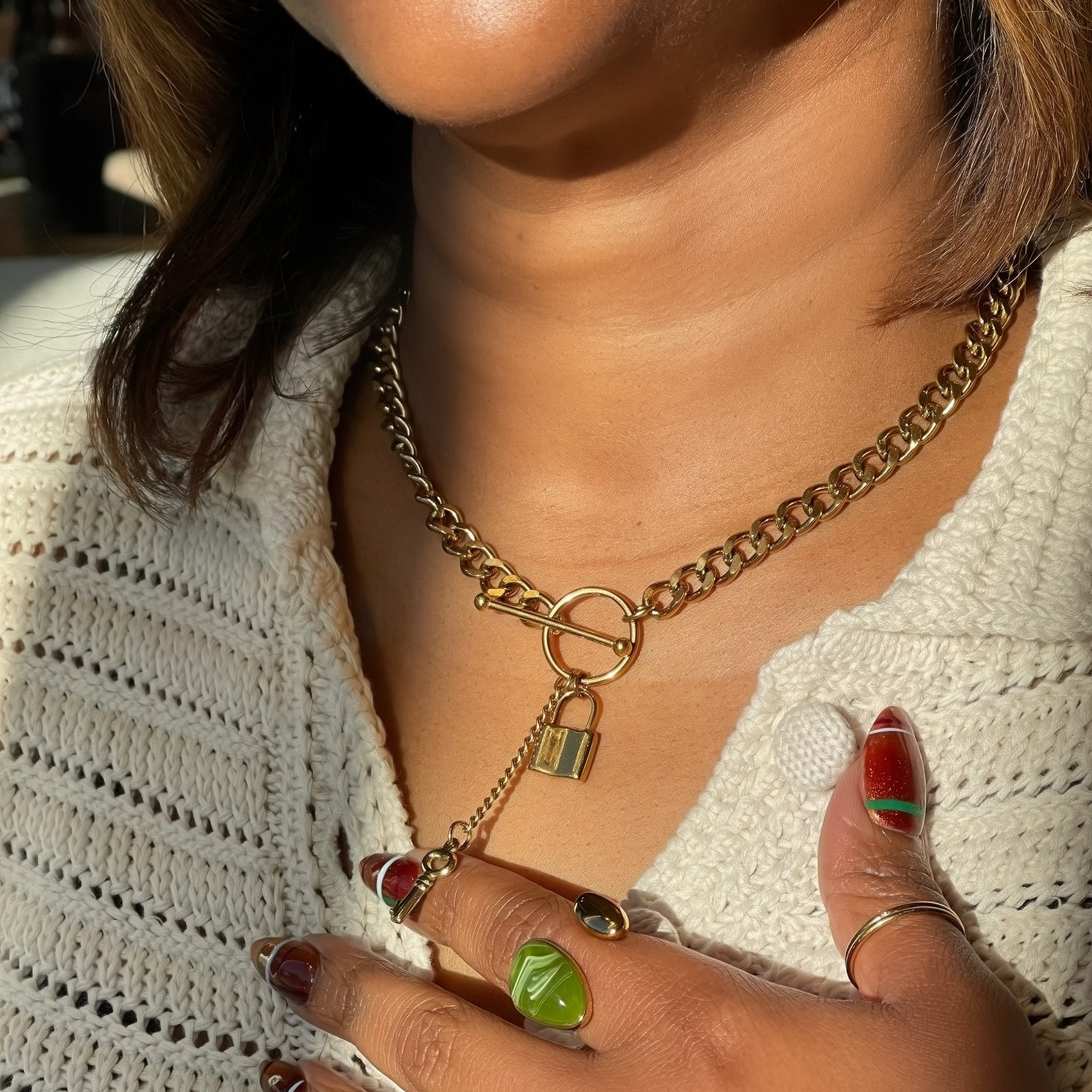 A close-up lifestyle shot of a person wearing a gold chain necklace with a padlock charm and a small drop chain with a green pendant, styled with a white knit top.