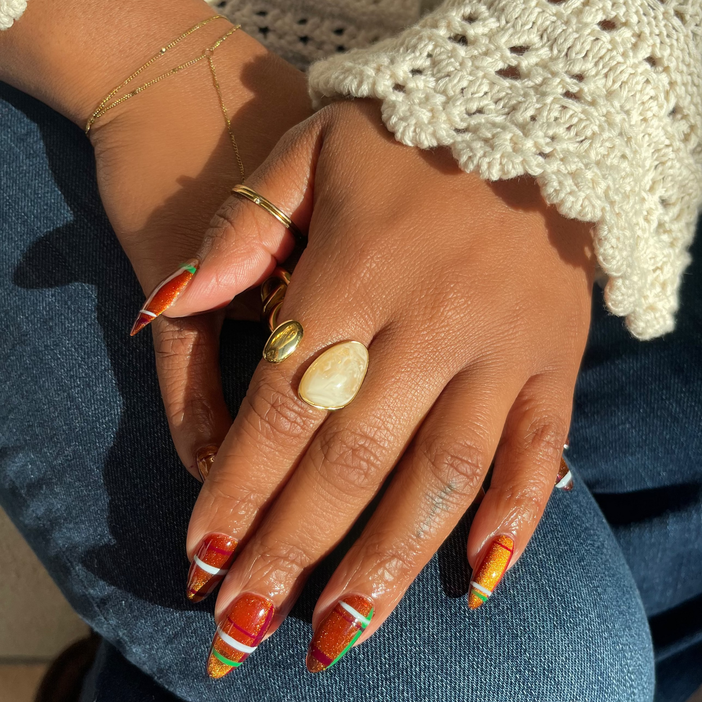 A close-up of a person's hands resting on their lap, showcasing a set of long stiletto nails painted with a colorful autumn pattern of orange, yellow, and green. The person is wearing a cream-colored crochet cardigan and blue jeans, with several gold rings on their fingers.