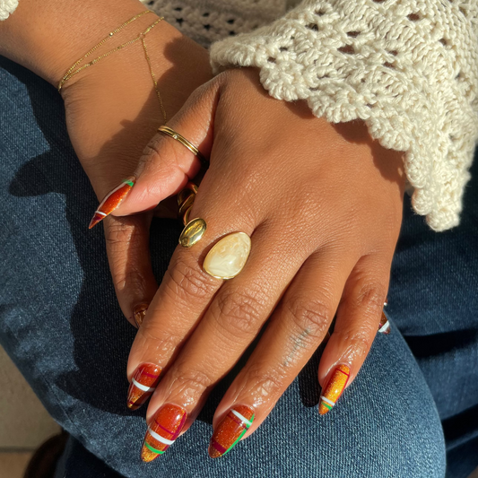 A close-up of a person's hands resting on their lap, showcasing a set of long stiletto nails painted with a colorful autumn pattern of orange, yellow, and green. The person is wearing a cream-colored crochet cardigan and blue jeans, with several gold rings on their fingers.