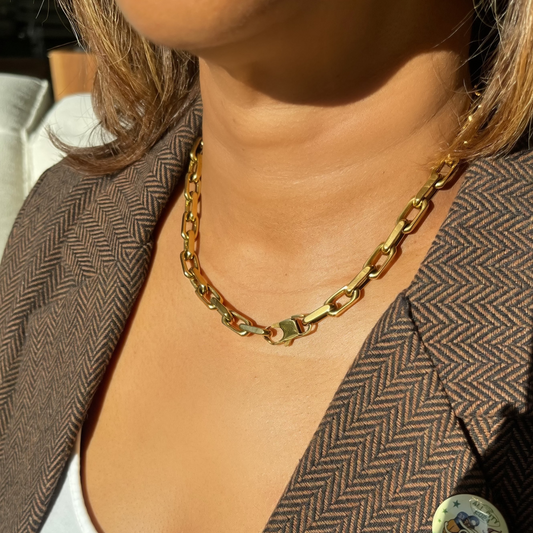 A close-up lifestyle shot of a woman wearing a chunky gold chain necklace with a lobster clasp, styled with a brown herringbone blazer and white top.