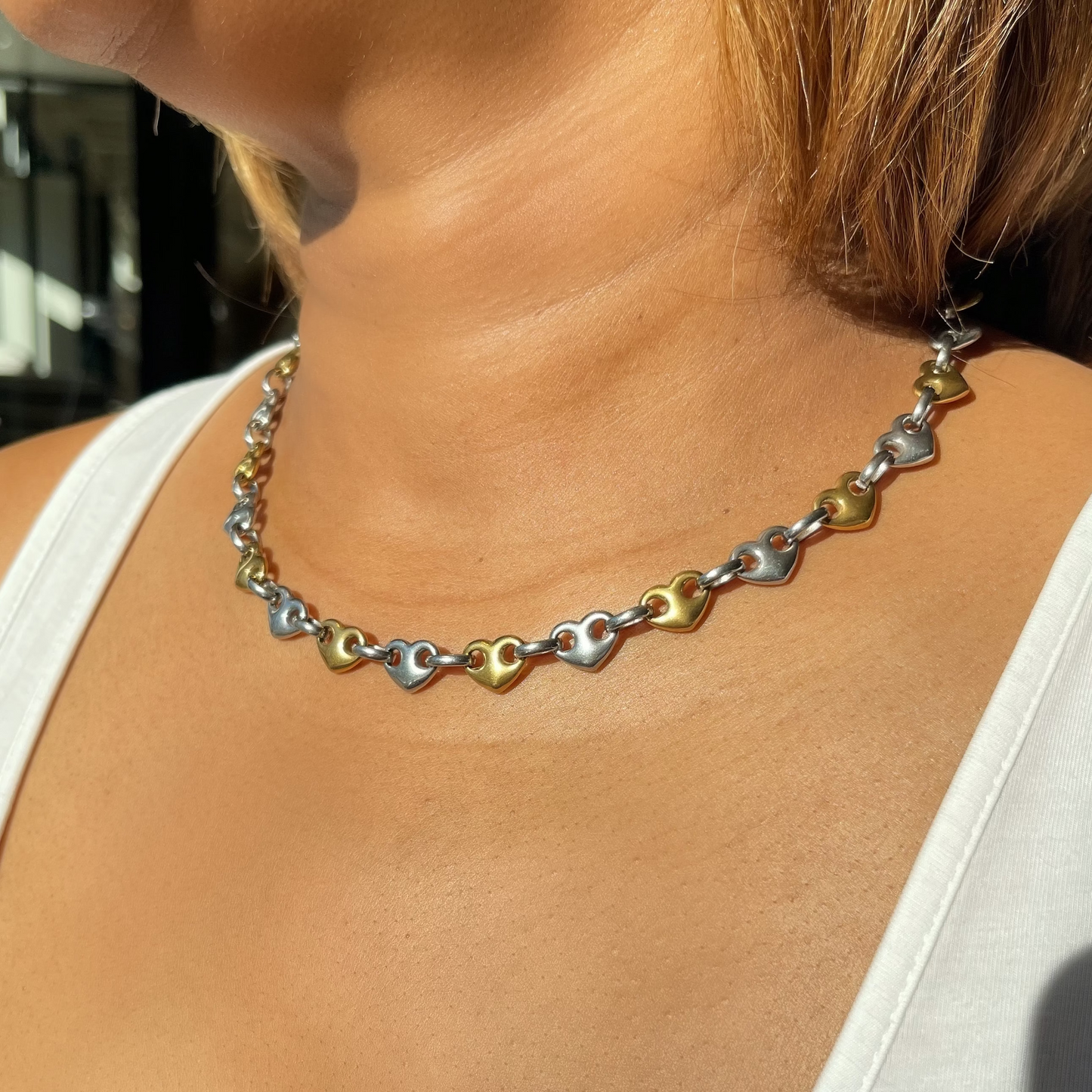 A close-up of a woman wearing a heart-link necklace with alternating gold and silver hearts, styled with a white tank top.