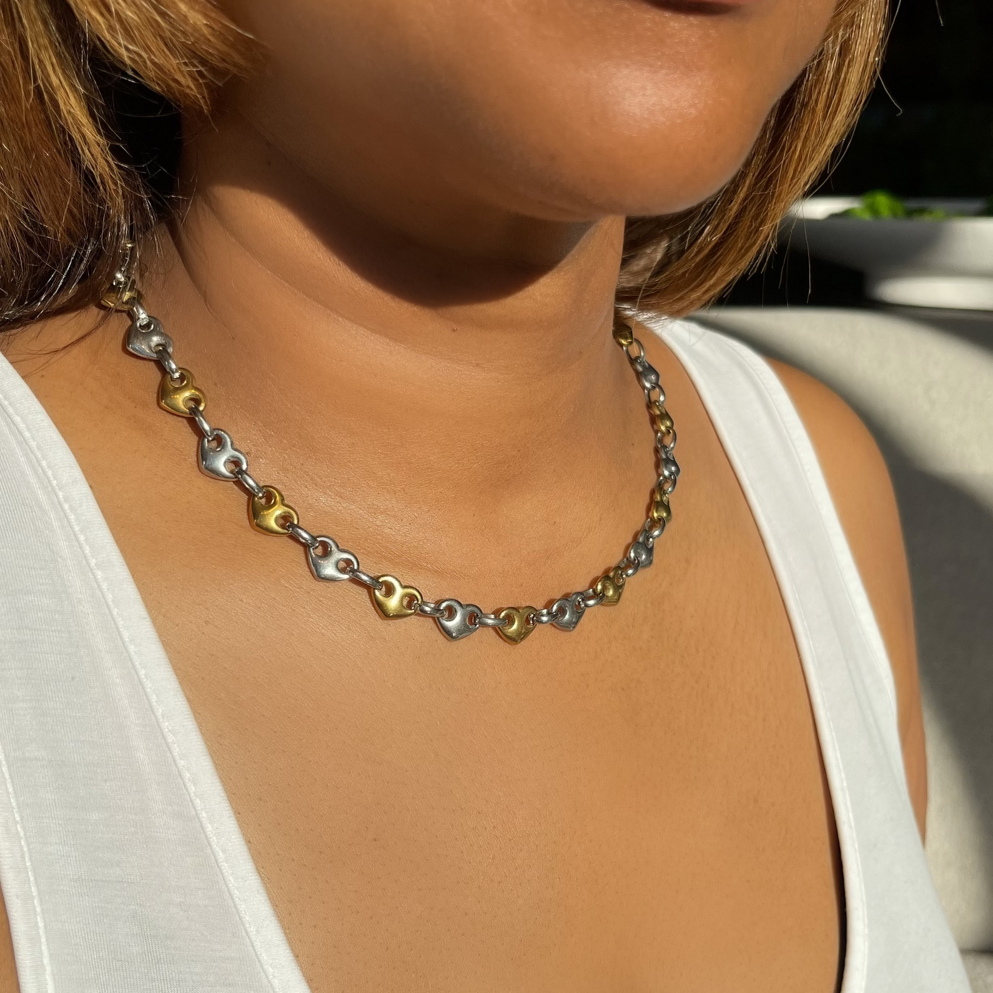 A close-up lifestyle shot of a woman wearing a delicate heart-link chain necklace with alternating silver and gold hearts, paired with a white tank top.