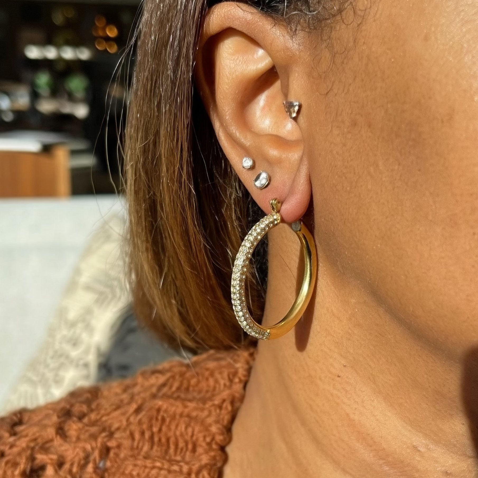 A close-up lifestyle shot of a person wearing a large gold hoop earring with a textured silver inner band, along with small studs and a cartilage piercing. The person is wearing a brown knit sweater and black-framed glasses, with warm sunlight casting shadows on their skin.