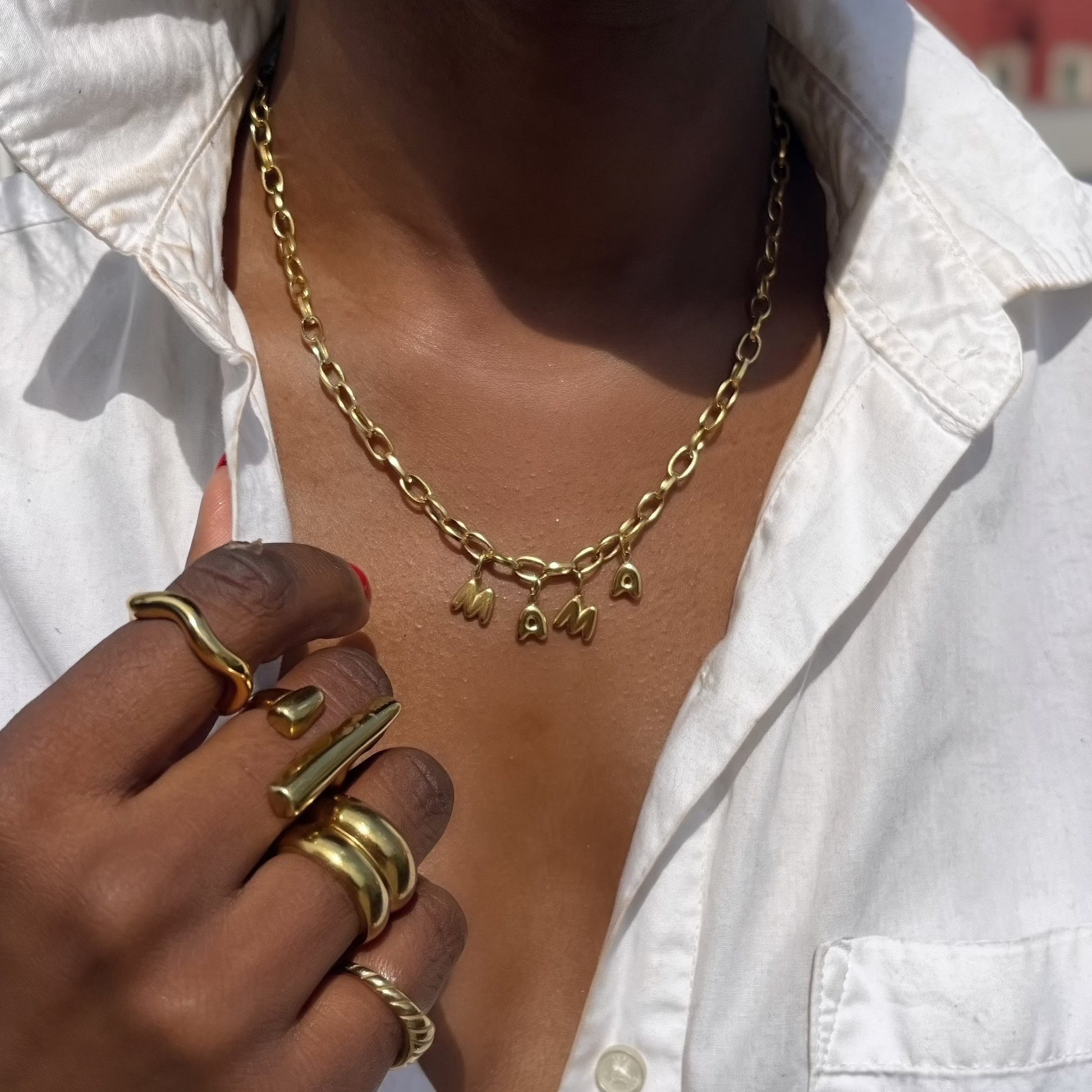 Person wearing a gold mama necklace and multiple rings on a blurred outdoor background