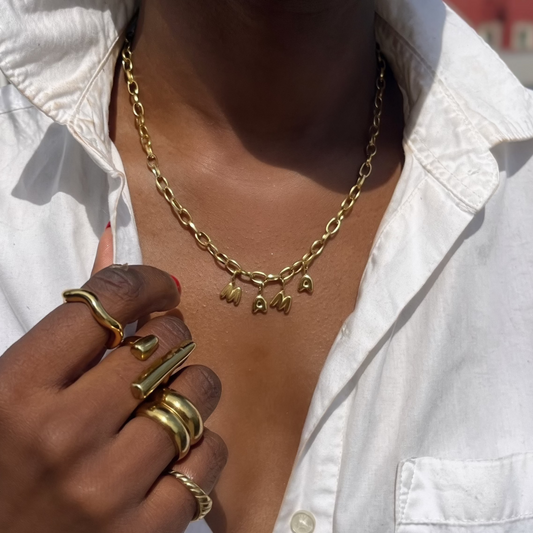 Person wearing a gold mama necklace and multiple rings on a blurred outdoor background
