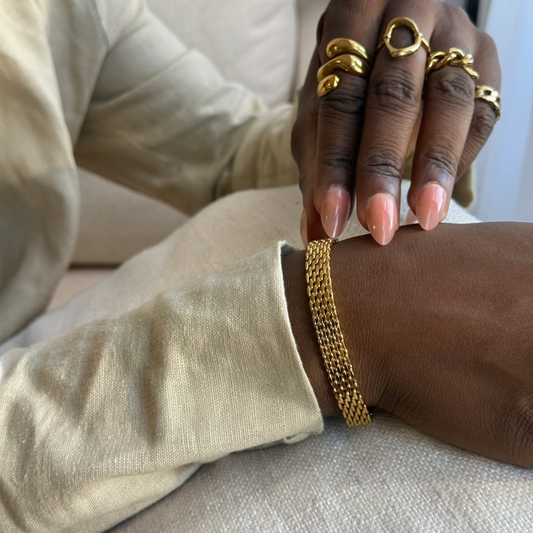 A close-up lifestyle shot of a person wearing a gold chain mesh bracelet on their wrist, with their hand resting on their knee. They are also wearing multiple gold rings on their fingers.