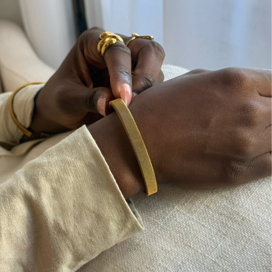A close-up lifestyle shot of a person wearing a thin gold mesh bracelet on their wrist, with their other hand adjusting or touching the bracelet. They are wearing a beige long-sleeve top and gold rings on their fingers, sitting on a light-colored sofa.
