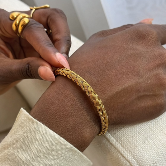 A close-up of a person's wrist wearing a gold braided chain bracelet, with their other hand adjusting or touching the bracelet. The person is also wearing a gold snake ring on one finger and a light beige long-sleeve shirt.