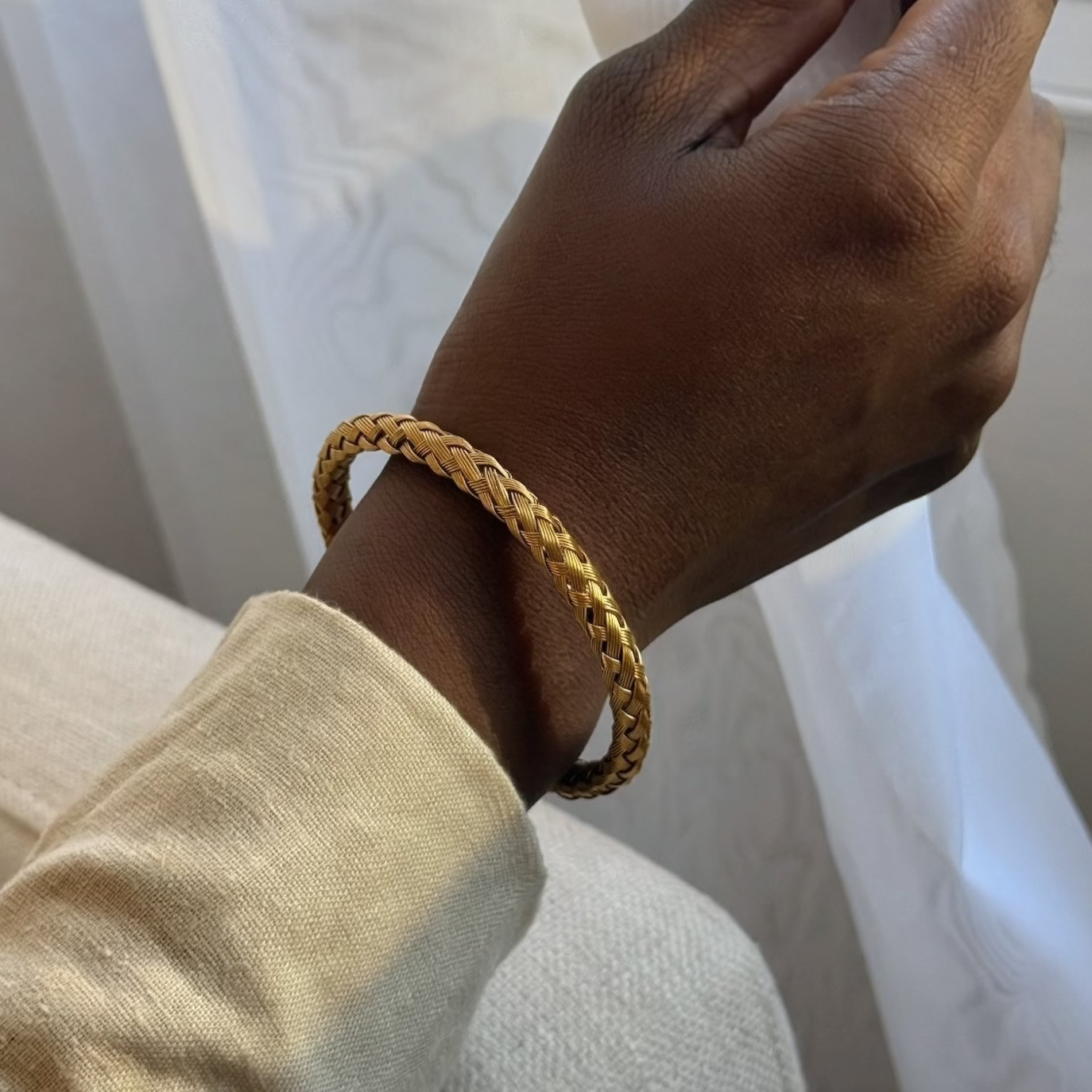 A close-up of a person's hand wearing a gold braided bangle bracelet, holding a white curtain in a softly lit indoor setting.