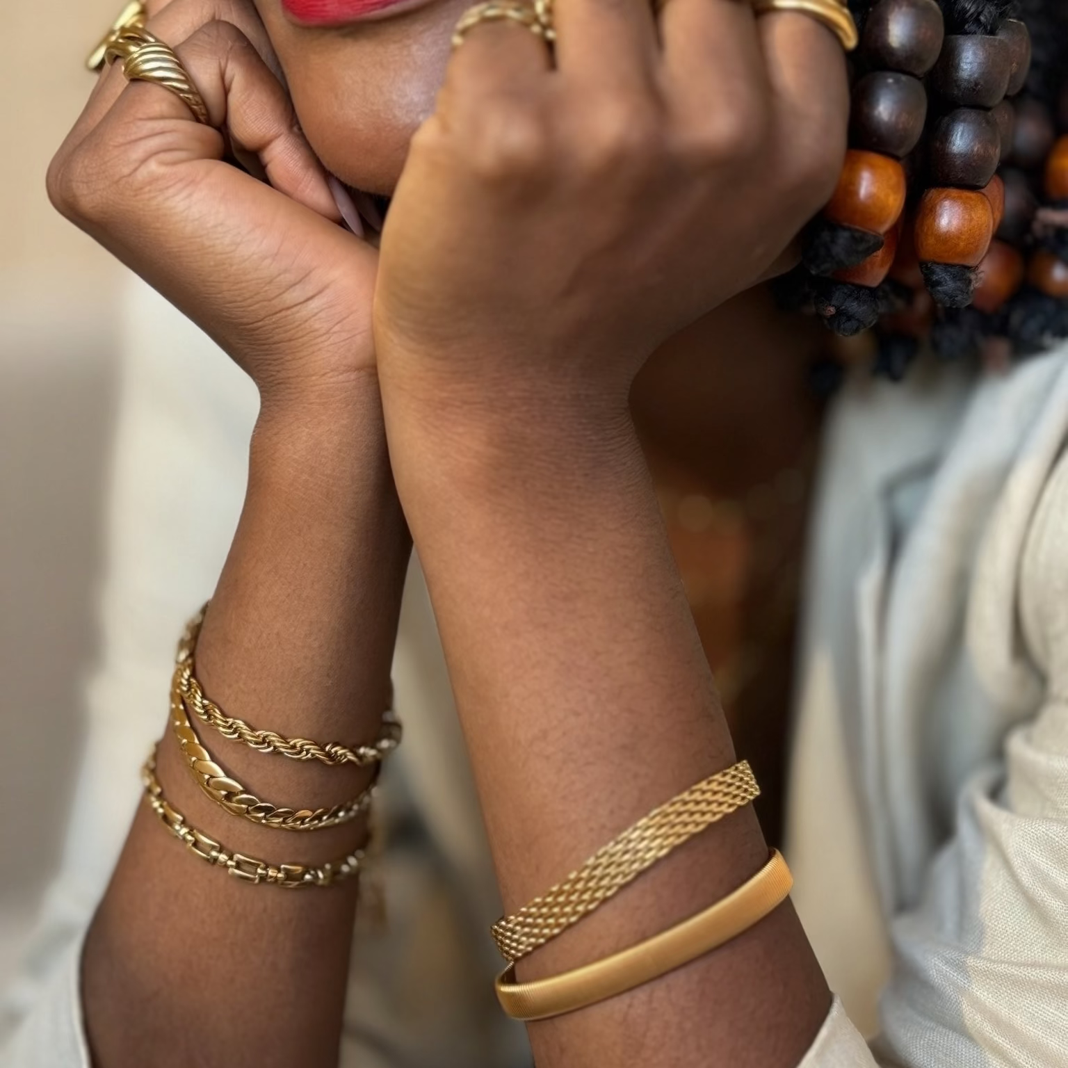 Close-up of a person wearing multiple gold-toned jewelry pieces, including stacked chain bracelets, a mesh bangle, and several rings, with red lipstick and braided hair visible.