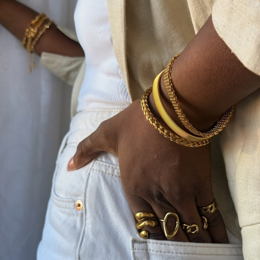 A close-up lifestyle shot of a person wearing a beige linen jacket, white jeans, and multiple gold bracelets and rings, with their hand in their pocket.