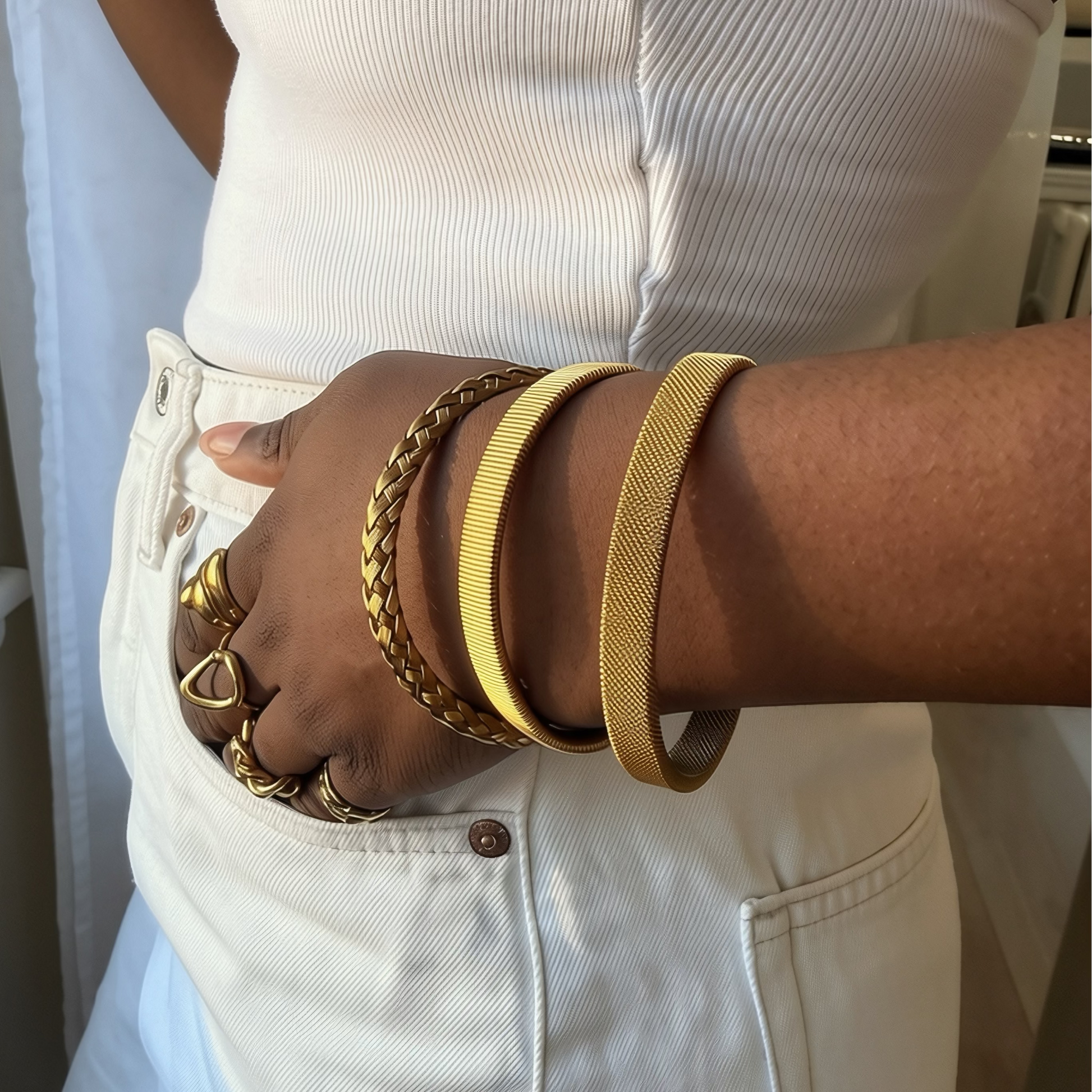 A close-up of a person wearing a white ribbed strapless top and white pants, with a hand in the pocket wearing multiple gold-toned bracelets and rings.