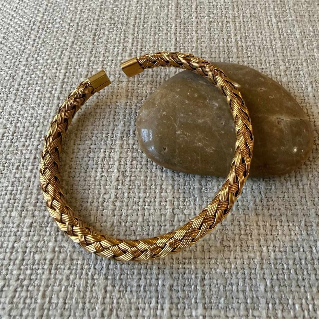 A braided gold-toned cuff bracelet resting on a textured beige fabric surface with two smooth stones behind it.