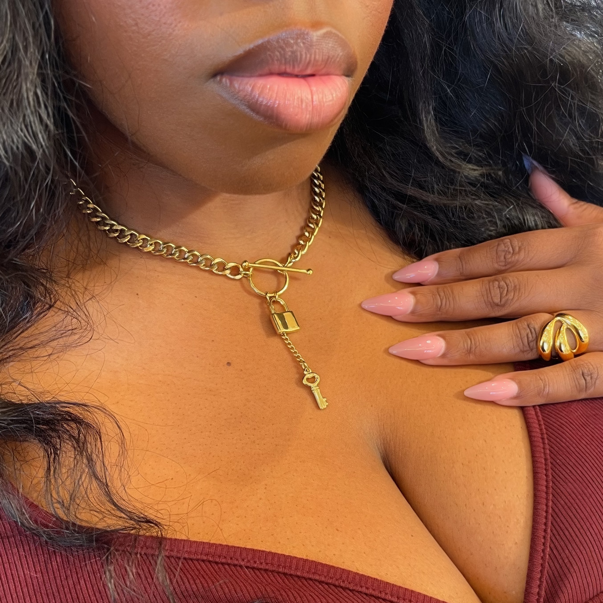 A close-up lifestyle shot of a woman wearing a gold chain necklace with a padlock and key charm, paired with a burgundy ribbed top.