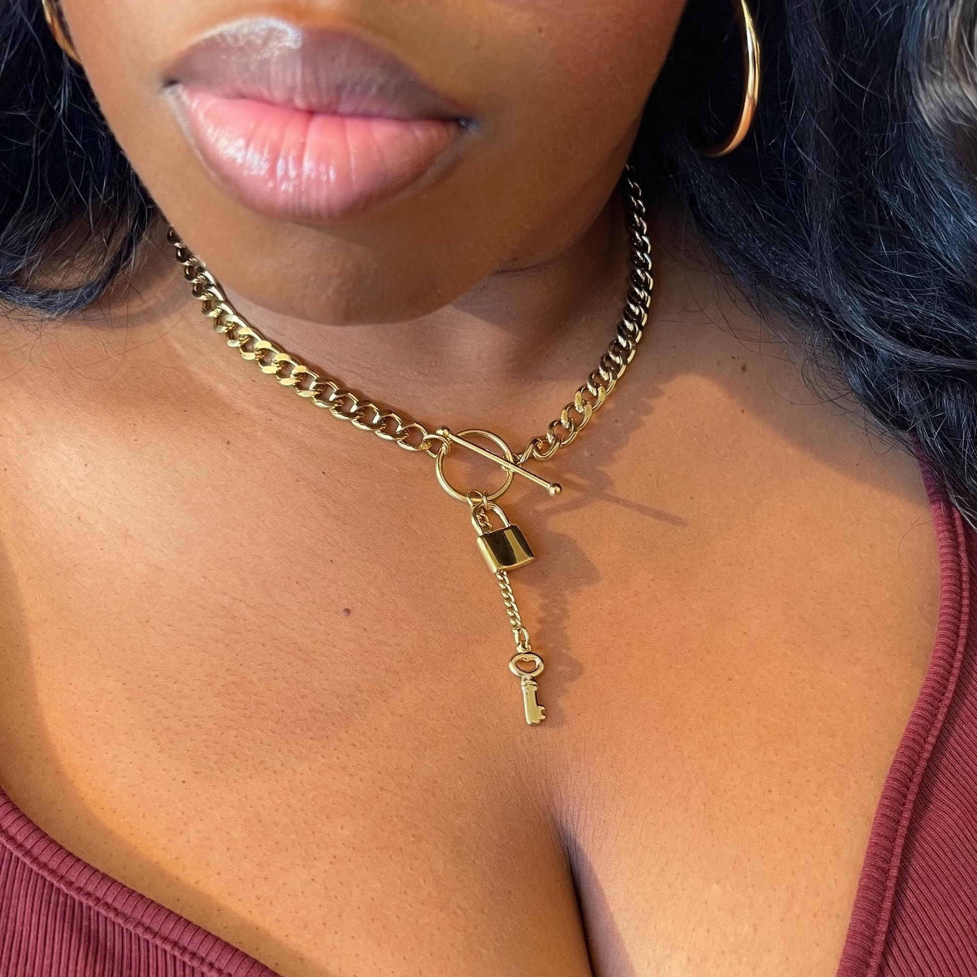 A close-up lifestyle shot of a woman wearing a gold chain necklace with a padlock and key pendant, paired with a burgundy ribbed top.