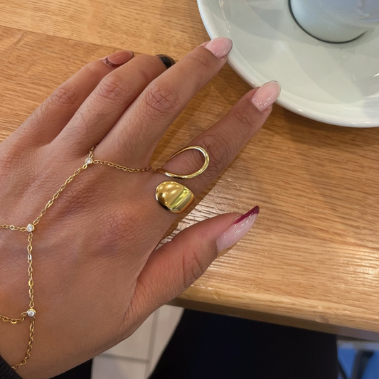 A close-up of a hand wearing a gold chain hand bracelet and two gold rings, resting on a wooden table next to a white coffee cup on a saucer.