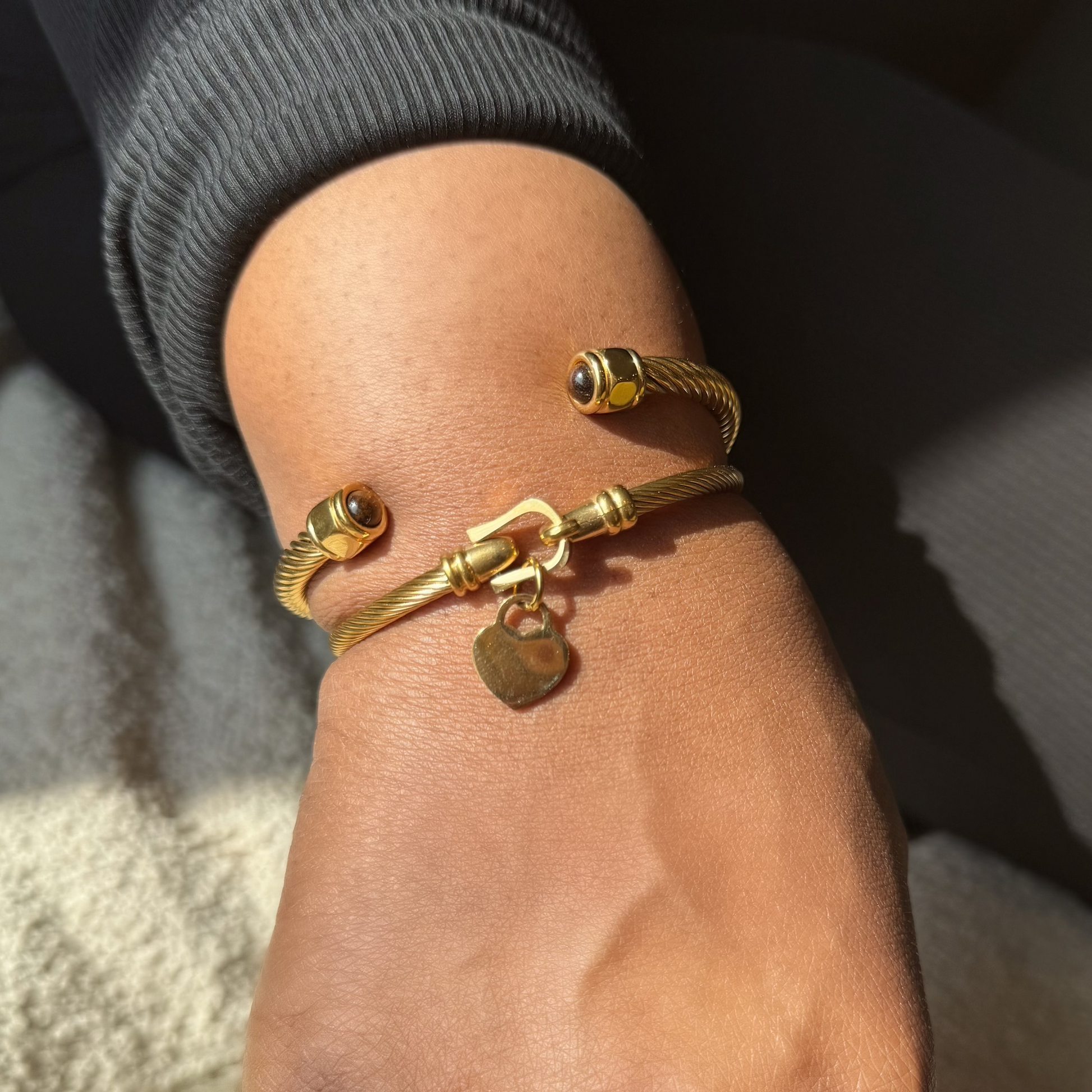 A close-up of a person's hand wearing a gold cuff bracelet with a small heart charm, along with a gold ring, set against a dark gray top and textured beige background.