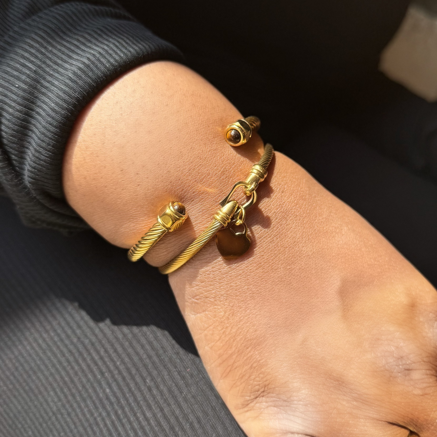 A close-up of a person's hand and wrist wearing three gold-toned bangle bracelets, including one with a small heart charm, along with matching gold rings on the fingers.