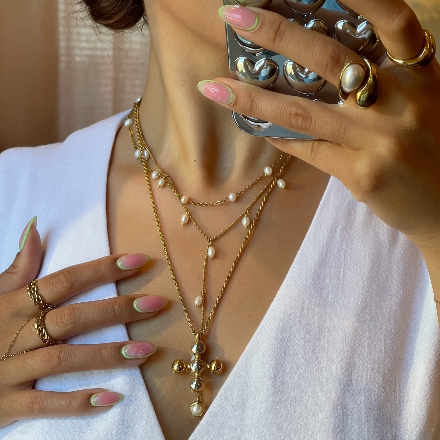 Person wearing a white outfit with gold jewelry, including necklaces and rings, holding a phone case with metallic design.