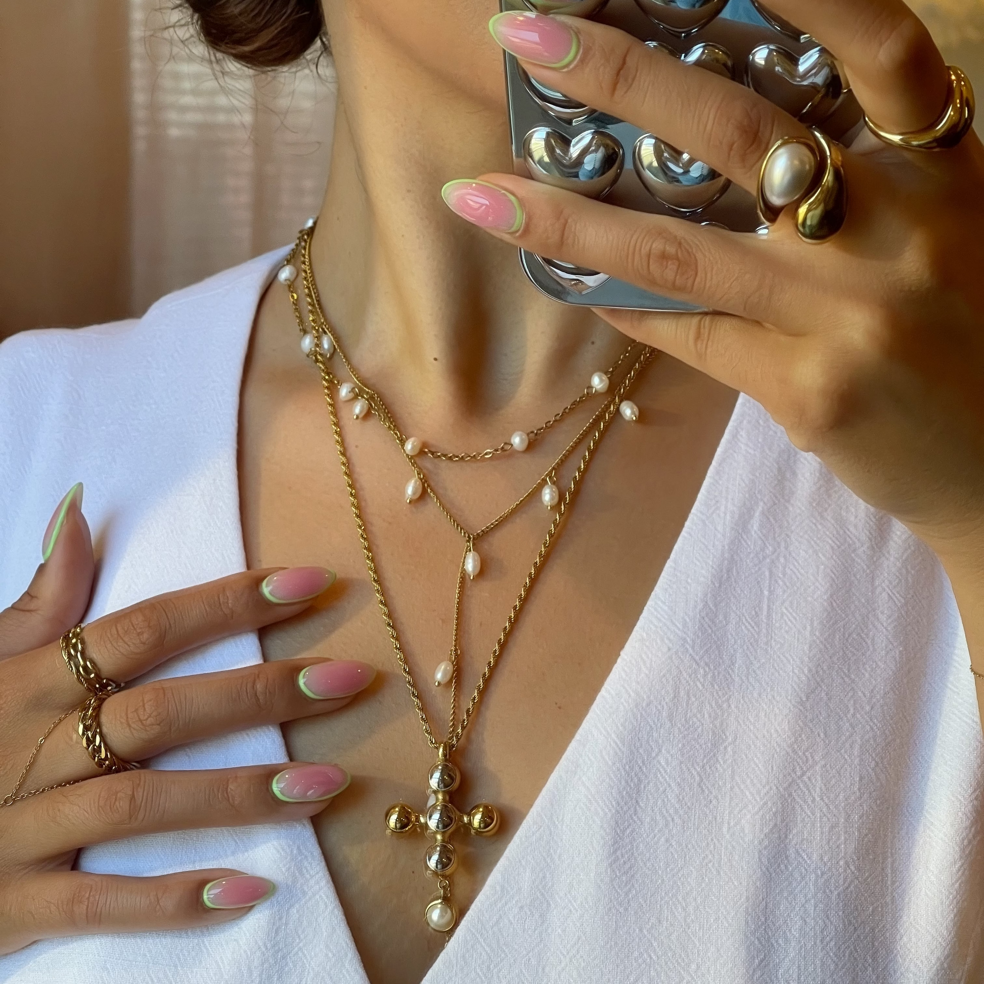 Person wearing a white outfit with gold jewelry, including necklaces and rings, holding a phone case with metallic design.