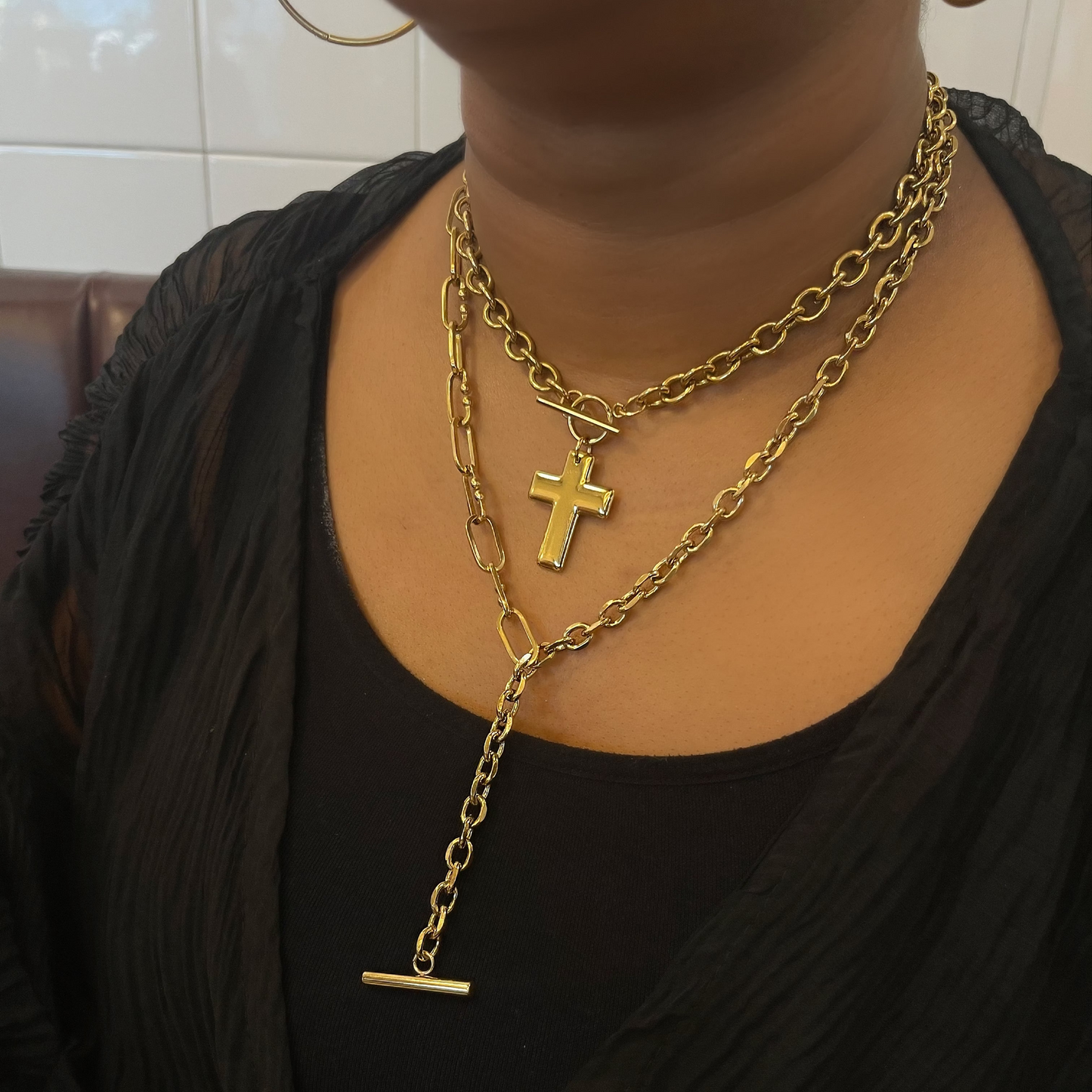 Gold chain necklace with a cross pendant worn by a person against a tiled wall background.