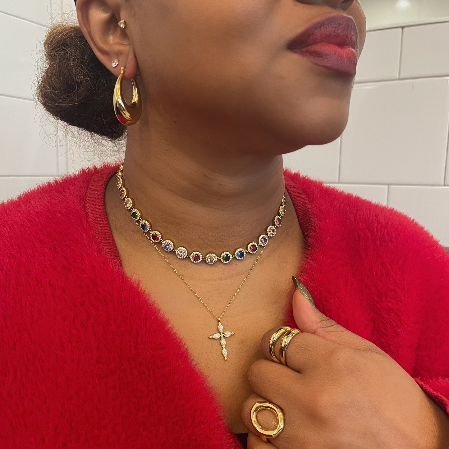 Woman wearing gold jewelry including earrings, necklace, and rings against a tiled wall background.