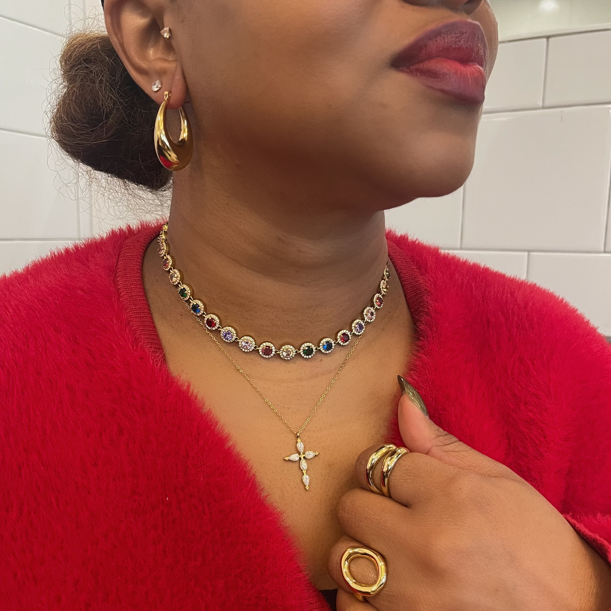 Woman wearing gold jewelry including earrings, necklace, and rings against a tiled wall background.