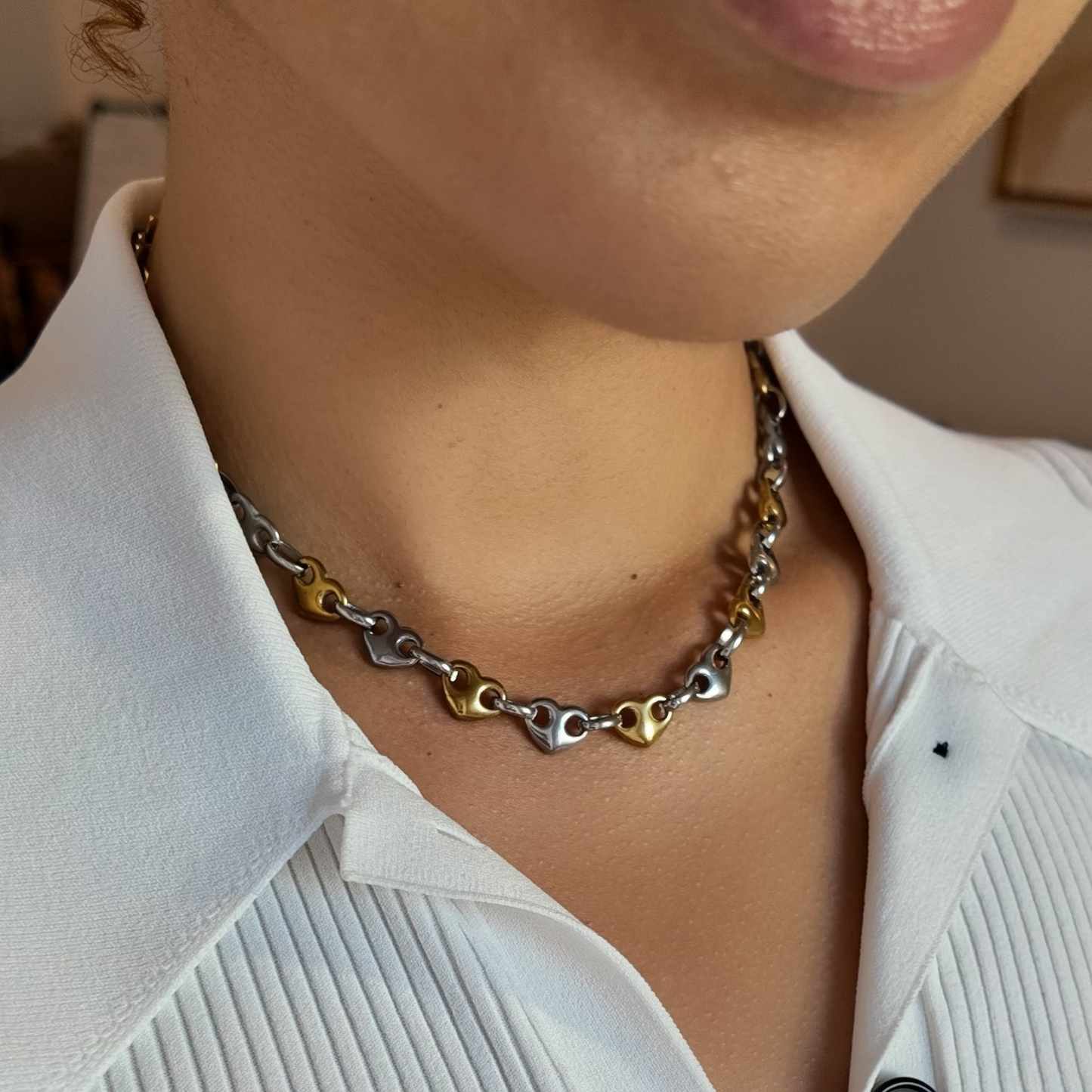 A close-up of a person wearing a two-tone metal chain necklace with interlocking heart links in silver and gold, styled with a white pleated button-up shirt.