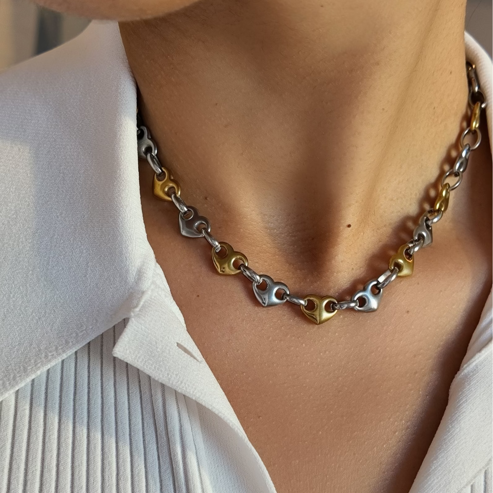 A close-up of a person wearing a multi-strand necklace with interlocking heart-shaped links in silver, gold, and rose gold tones, styled with a white pleated collared shirt.