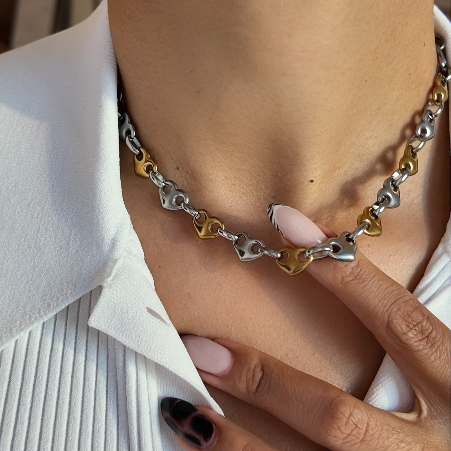 A close-up lifestyle shot of a person wearing a silver and gold heart-link chain necklace, styled with a white pleated collared shirt.