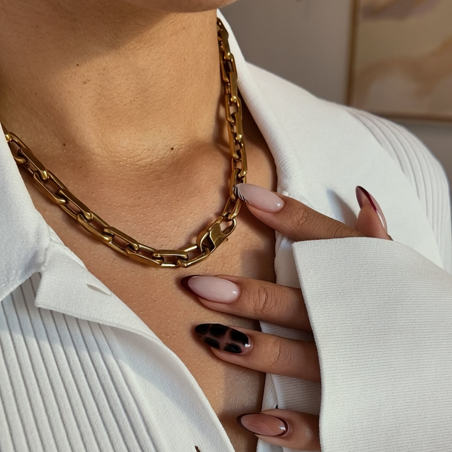 A close-up lifestyle shot of a person wearing a chunky gold chain necklace over a white pleated button-up shirt. The person's hand with manicured nails rests on their chest.