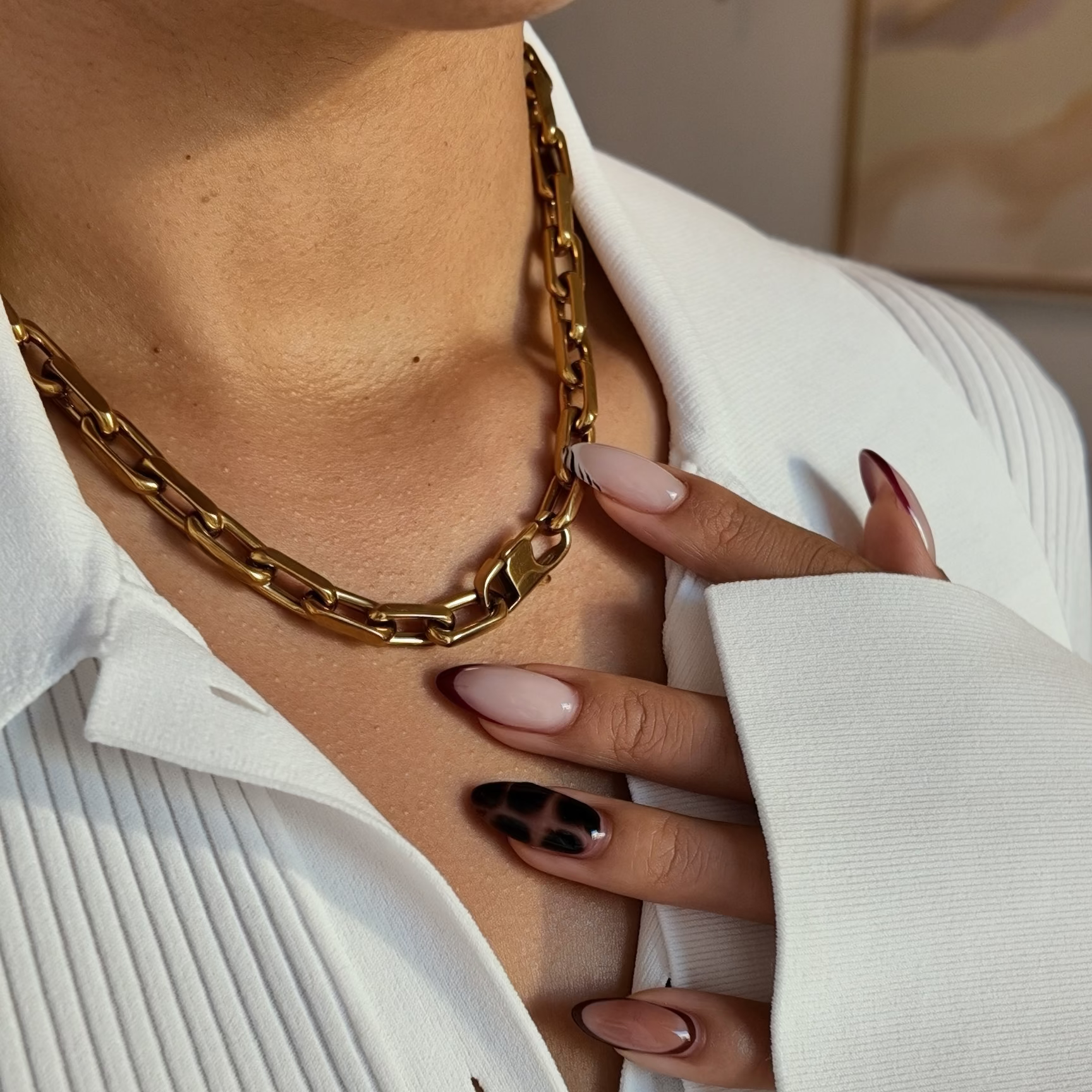 A close-up lifestyle shot of a person wearing a chunky gold chain necklace over a white pleated button-up shirt. The person's hand with manicured nails rests on their chest.