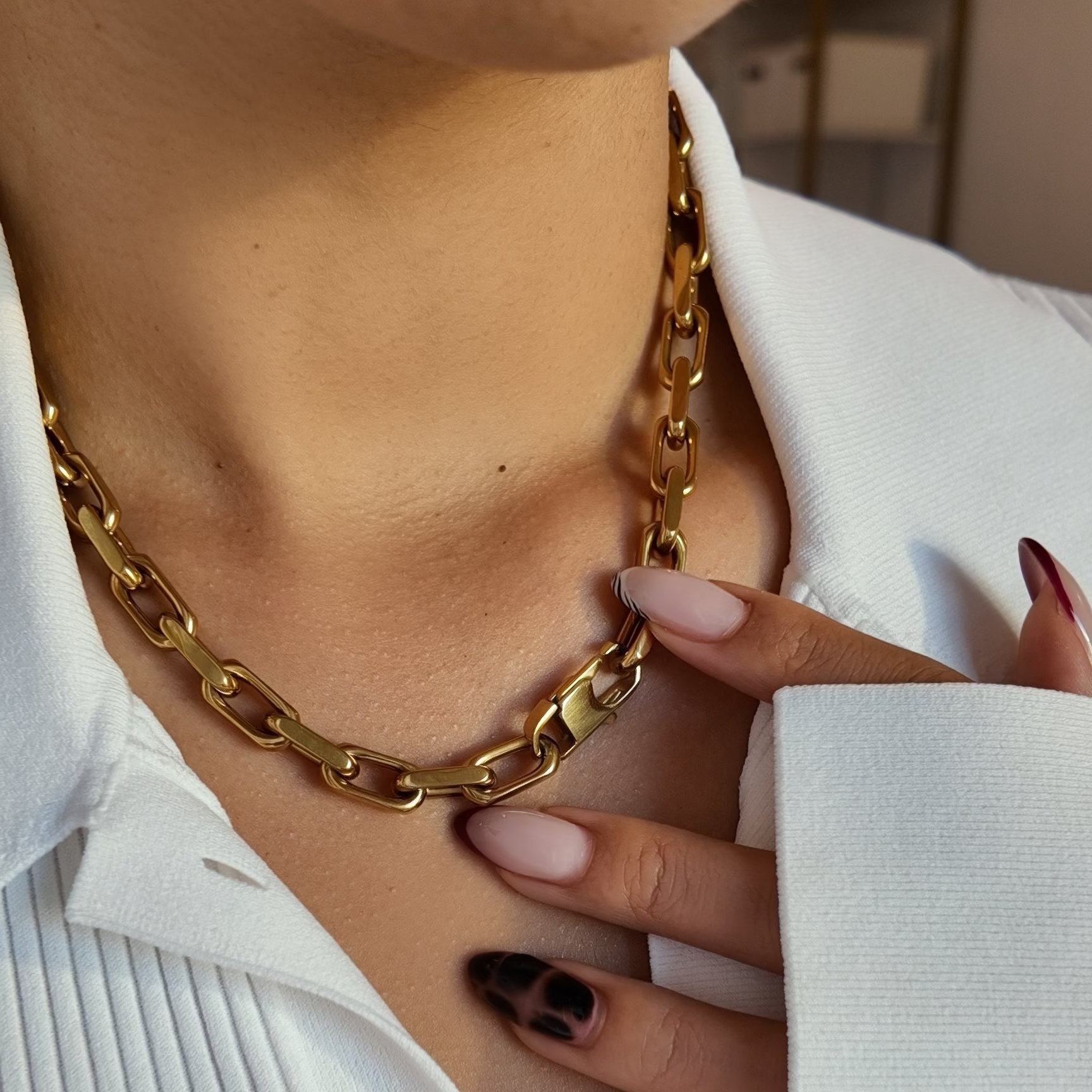 A close-up of a person wearing a chunky gold chain necklace over a white pleated collared shirt, with one hand touching the necklace.