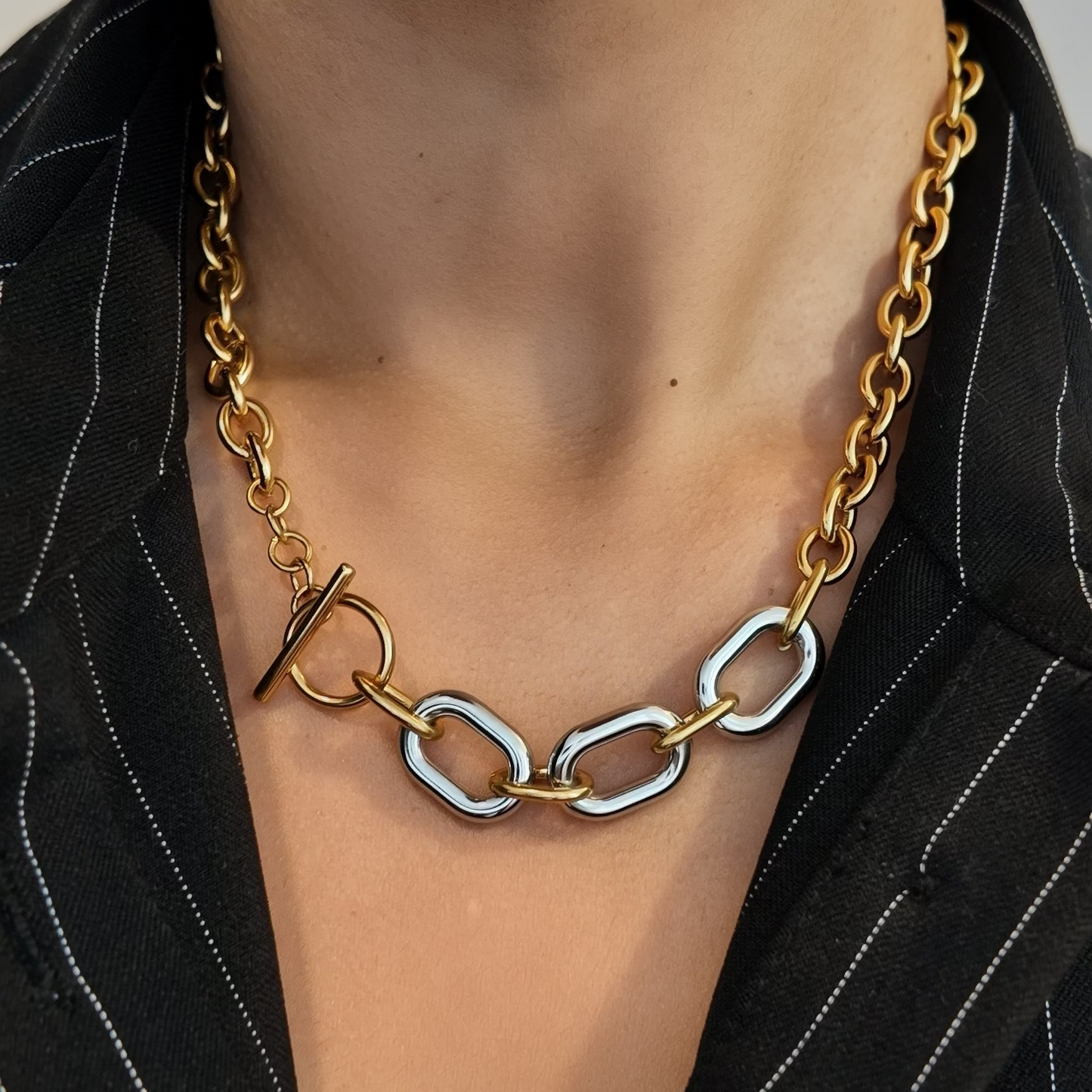 A close-up of a person wearing a chunky gold chain necklace with a toggle clasp and three large interlocking silver oval links, styled with a black pinstripe blazer.